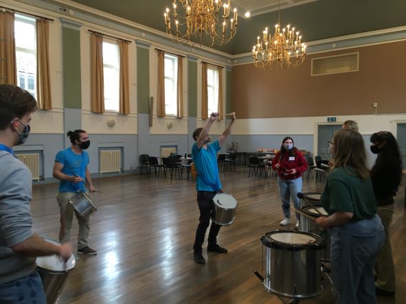Students stood in a circle playing drums in a large hall, as part of the African drumming workshop on field trip in Glastonbury Town Hall