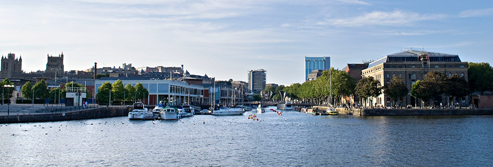 Bristol harborside in the sun, with the cathedral on the left and boats docked in the foreground.