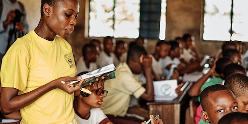 Busy classroom of students with a young Nigerian girl in the foreground stood up reading from a book.