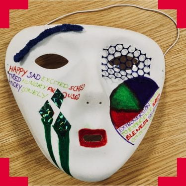 A photograph of a white mask with various drawings and materials adorning it. There is mesh over one eye, red around the mouth, felt on the forehead, and a written list of emotions on the left cheek