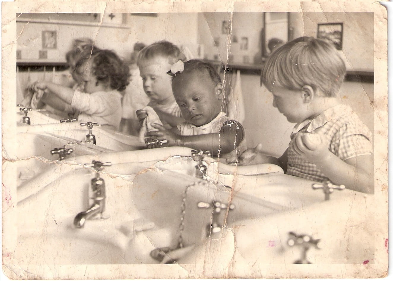 An old sepia photo of children washing up at a row of basins, the child in the centre of the photograph is a young black child, the other children are all white with one boy staring at the black child