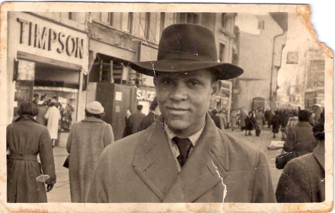 A sepia photo of a young male person of colour on what seems to be an English highstreet in the 1960s 