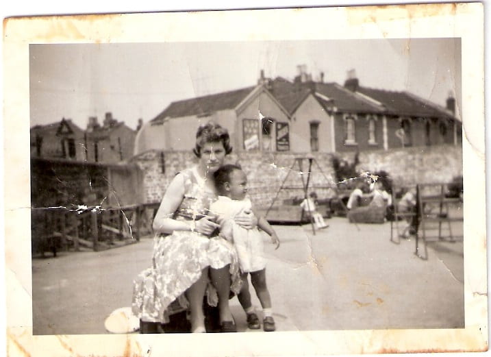 A sepia photograph of a white woman sat on a chair in the street holding a small black girl 