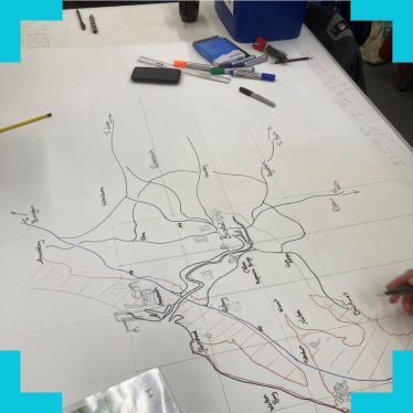 A photograph of a map on a school table with pens and other stationary scattered on it and students pointing to the map out of frame