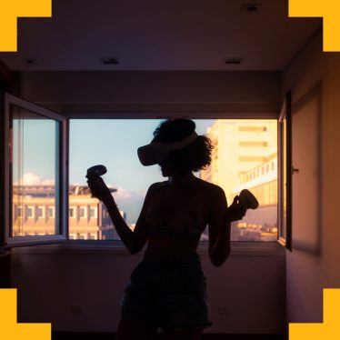 A photo of a woman stood in a room using a VR headset in front of an open highrise window 