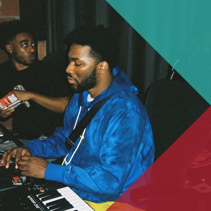 A group of young black men sat around a keyboard, a laptop, and music production equipment