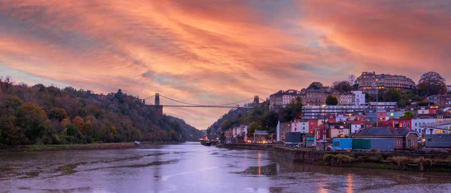 A view of the Clifton Suspension Bridge above Bristol harbour, at dusk with a dramatic orange sky