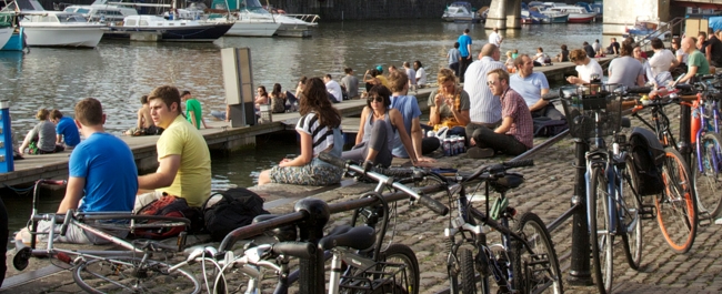 A large number of people sat along the waterfront at Bristol's Harbourside. There are several bikes attached to railings in the foreground, and several boats in the dock in the background.