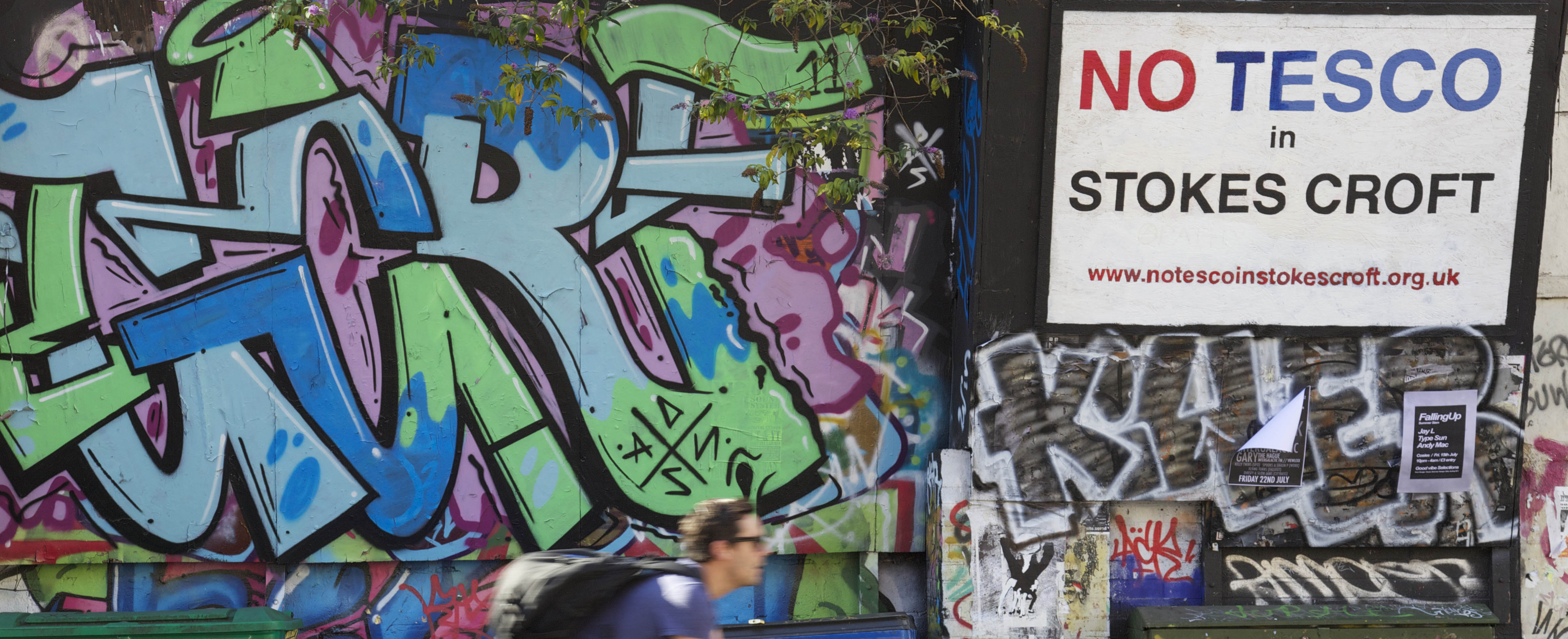 A graffiti'd wall in Stokes Croft with a sign that reads 'no tesco in stokes croft'. A man is cycling past.