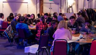 PhD students sitting at numerous round tables in a large room having discussions about PhD challenges 