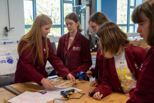 5 female sixth-form students from KLB school gather round a table to build their model aeroplane wing using wood, paper and hot glue