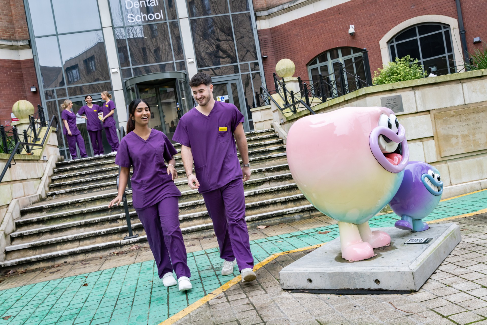 Two students in purple scrubs walking outside the Bristol Dental School building. A colourful statue of a tooth designed by Aardman Animation Studios is on the right.