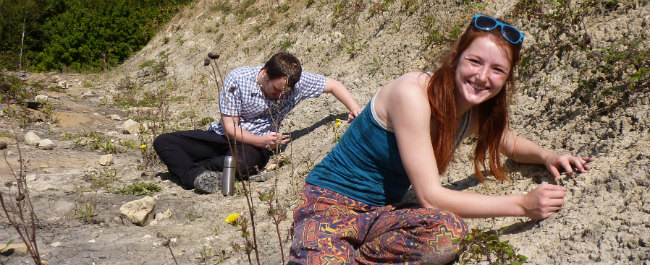 Two students sit on the ground on a field site, inspecting the rocky ground on a sunny day. The female student in the foreground of the image smiles broadly at the camera.