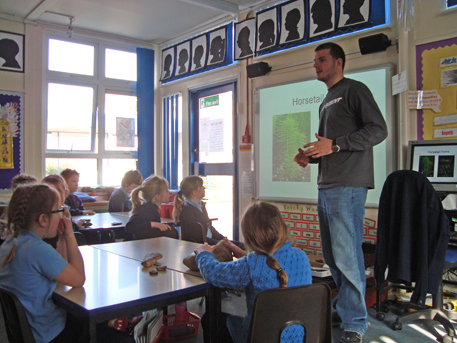A man stands at the front of a classroom, talking to an eager class of young Key Stage 2 children