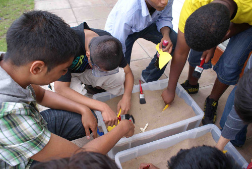 A group of school children are sat around a fossil and using tools to dig out a fossil.