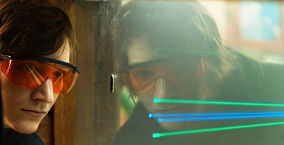 Female researcher in safety goggles reflected in screen with converging blue and green lines