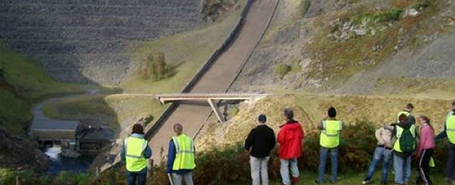 Researchers, some in high vis, looking at footbridge over road from side of hill