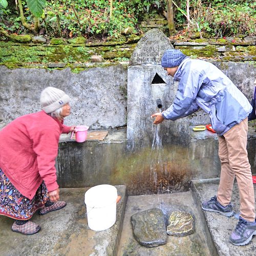 Two people collecting water from a tap which is fed by a traditional spring source in Nepal.