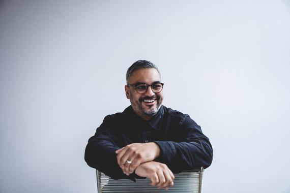 headshot of writer Nikesh Shukla - a middle aged man of Asian decent wearing glasses sitting on a chair and smiling