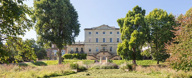 A large open green space with trees and a large white building towards the back