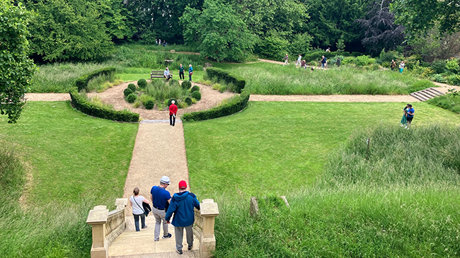 A large green space with a path going through the middle. There are a set of stone stairs leading down to the green space, with people walking down them. In the middle of the green space, there is a circular set of small hedges with people stood talking nearby