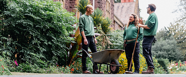 Three people stood up, facing each other talking. One of them is holding a broom and one is holding a wheelbarrow. They are all in green jumpers and black trousers.