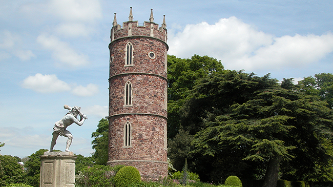 A tall thing bricked circular tower next to a white stone statue of a man holding an item behind his head. In the background are green trees and cloudy blue skies.
