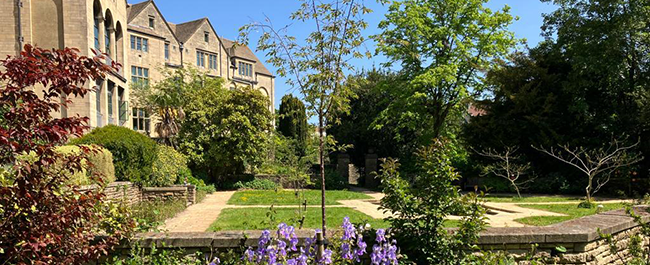 a small green space, surrounded by colourful flowers and green hedges. To the left of the garden are 4 old buildings with blue skies above.