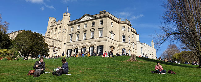 A large old building at the top of a small hill, with lush green grass and trees around it. Small groups of people are sat on the grass.