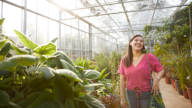 A person laughing, looking to the side of the camera. They are inside a large greenhouse which is filled with variety of green plants
