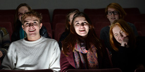 Male and female students sat in a cinema, gazing directly to the front, smiling and laughing as they watch a film.
