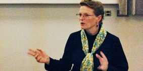 A middle-aged woman wearing glasses, dressed in a sweater and scarf talking and gesturing to an audience. 