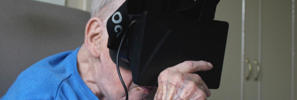 An elderly man is sitting down, holding a VR headset to his face - it covers his eyes and nose.