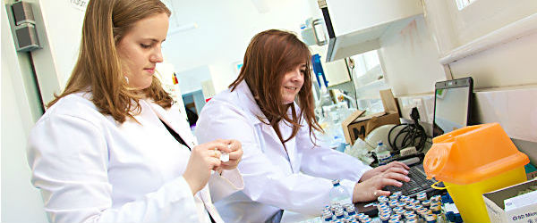 Two women wearing white coats in a laboratory. There are vials and a laptop on the table in front of them.