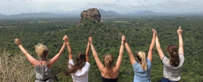 A group of girls with their arms in the air in celebration, look out at the view from the top of the mountain.