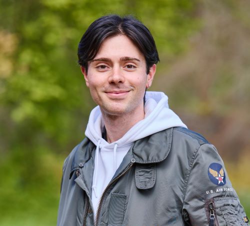 A student with short black hair parted in the middle. They are wearing a grey hoodie and a grey army style jacket. The blackground is blurred greenery.