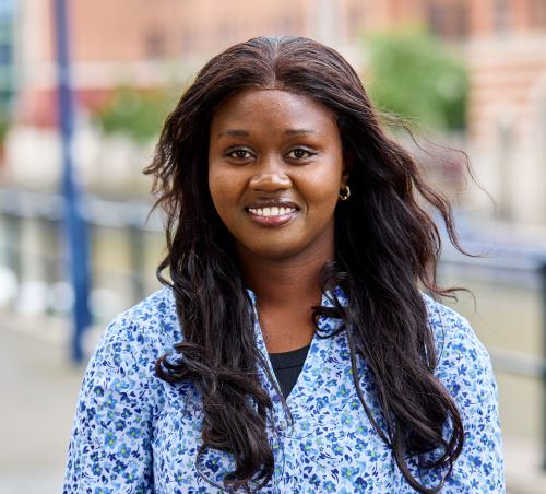 A student with long dark red hair wearing a blue and white patterned shirt. The background is a blurred image of a path by a waterway.