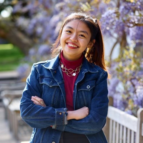 A student with long brown hair in a ponytail and their arms folded. They are wearing a red polo neck and a dark denim jacket. They are wearing gold earrings and a charm necklace.