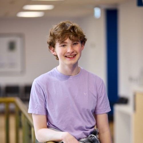 A student with blonde short hair leaning against a wood and glass railing. They are wearing a purple t shirt.