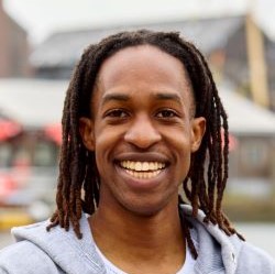 A student with shoulder length locs. They are wearing a grey hoodie and a white t shirt. The background is a blurred image of the Harbourside.