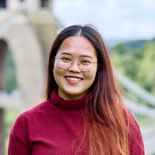 A student with long dark hair that is highlighted towards the bottom. They are wearing a red long sleeve top and glasses. A blurry image of a bridge is in the background.