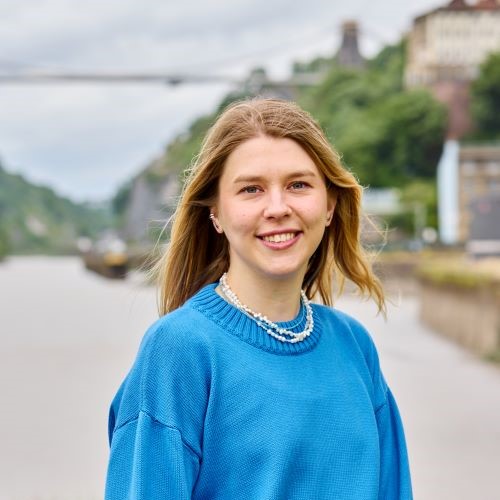 A student with long blonde hair that is blowing in the wind. They are wearing a bright blue jumper, a beaded necklace, and a small silver cuff in their ear. Behind them is a blurred background of the Clifton Suspension bridge and the Avon Gorge..