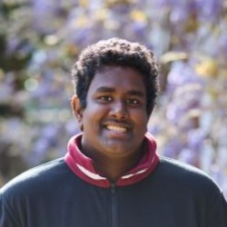 A student with dark black hair wearing a black fleece with a white and red striped collar. The background is blurred wisteria.