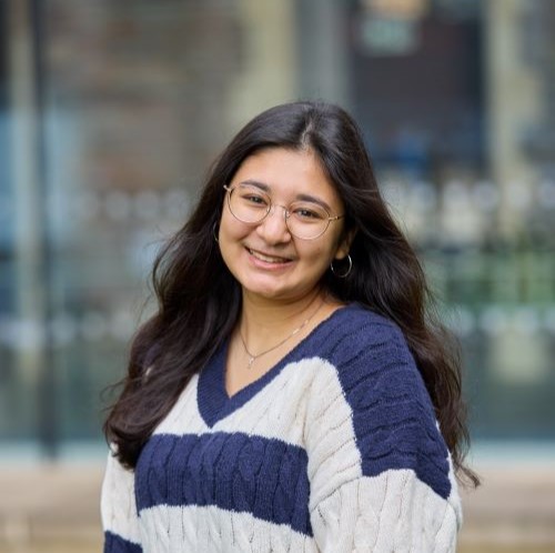 A student with long dark hair. They are wearing a blue and white striped jumper, silver glasses, silver hoops and a silver necklace. The background behind them is a blurred glass wall.