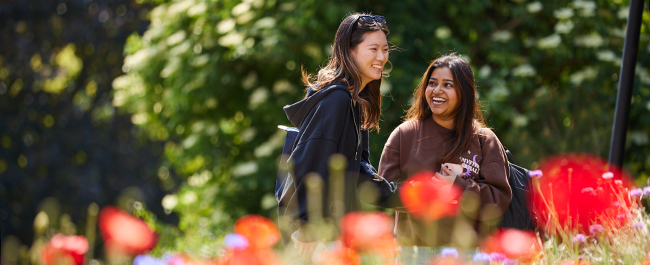 Two female students laughing, with greenery in the background and colourful flowers out of focus in the foreground.
