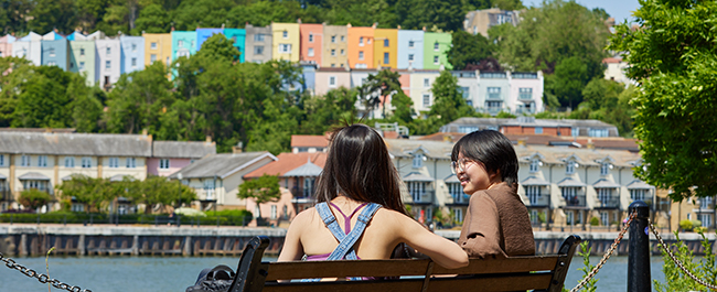 Two students sitting on a bench looking out onto the harbour and rows of coloured houses.
