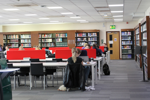 students working on rows of desks, the wall behind them is covered in bookshelves. 