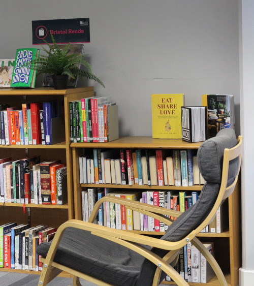 A reclining chair in front of two bookcases full of books, on top of one is a fern in a plant pot. The Bristol Reads sign is hung above the shelves.