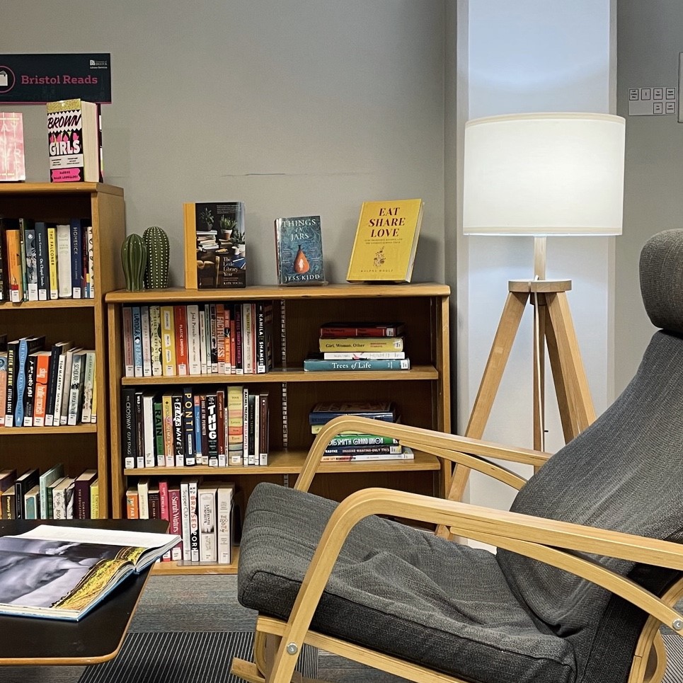 Bristol Reads area at the Arts and Social Sciences Library. Light wooden bookcases with books, next to a modern wooden lamp and Scandinavian-style armchair, also made from light wood.
