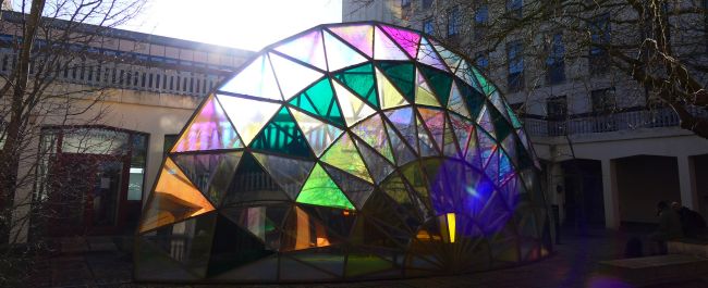 A large fan shaped sculpture full of different coloured mosaic pieces which the sun is shining through. This artwork is called 'Palm Temple', and is outside the Chemistry library.